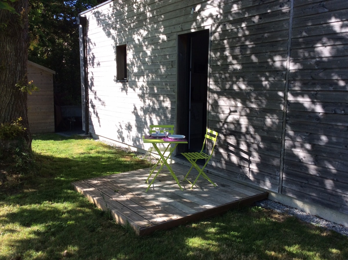 An outdoor terrace features a wooden platform with two green chairs and a small table, set against the backdrop of a wooden building. Dappled sunlight creates shadow patterns on the grass surrounding the area, enhancing the natural setting.