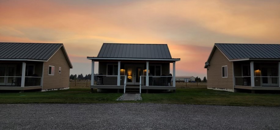 A row of newly built cabins, each featuring a covered porch and large windows. A gravel path leads to the cabins, set against a colorful sunset sky. The landscape is open and spacious, with lawns extending toward the horizon.