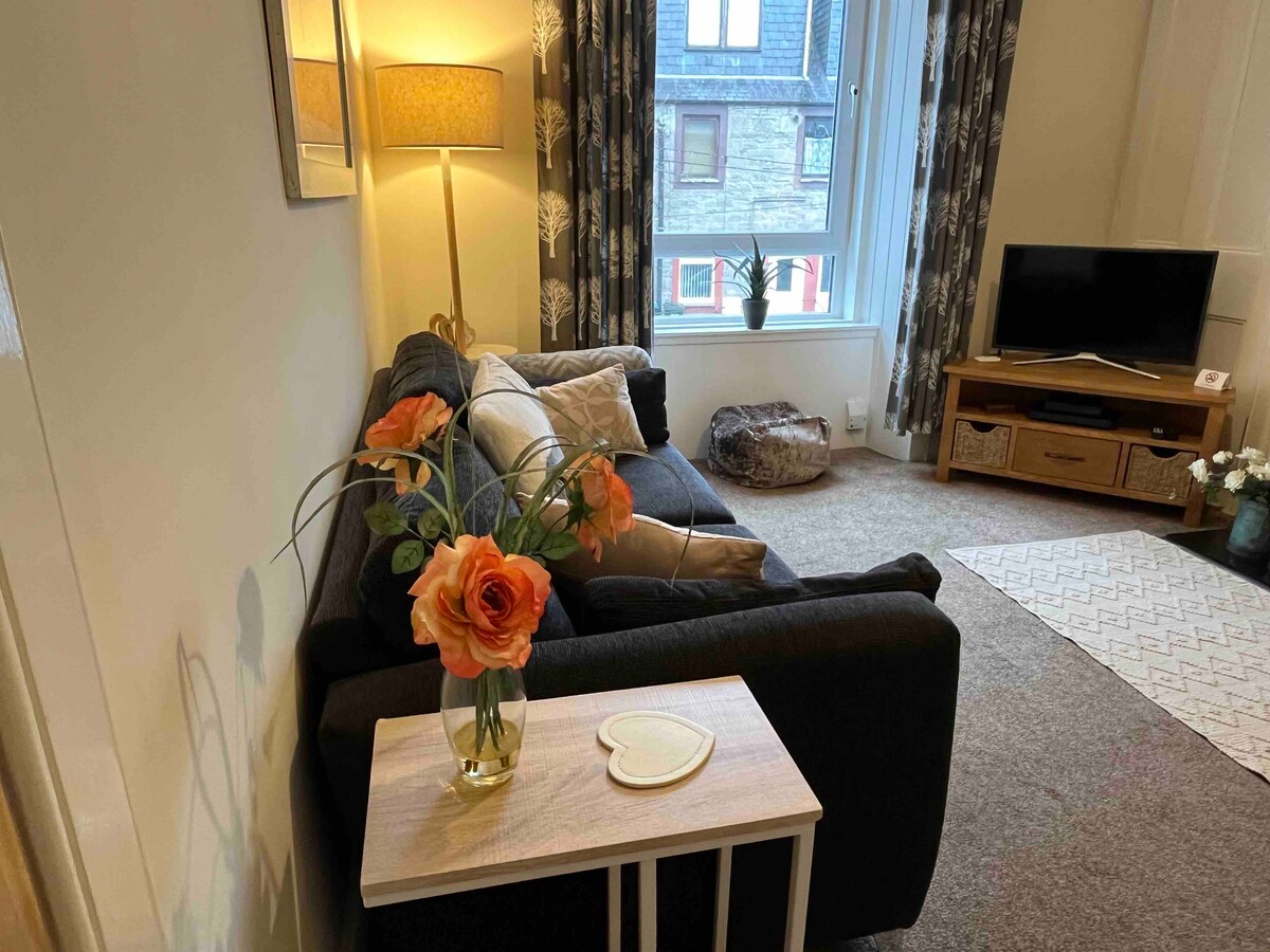 A cozy lounge area is depicted, featuring a dark sofa accented with decorative pillows. A side table holds a vase with orange flowers. Natural light filters through a window adorned with patterned curtains, illuminating a Smart TV and wooden media console.