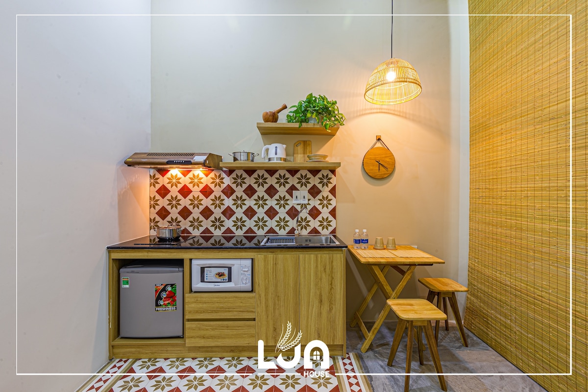 The kitchen area features eco-friendly wooden cabinetry, equipped with modern appliances including a refrigerator and stove. A decorative tile backsplash adds a distinctive touch. A small dining table with two wooden chairs is positioned nearby, complemented by a pendant light and greenery on the shelf above.