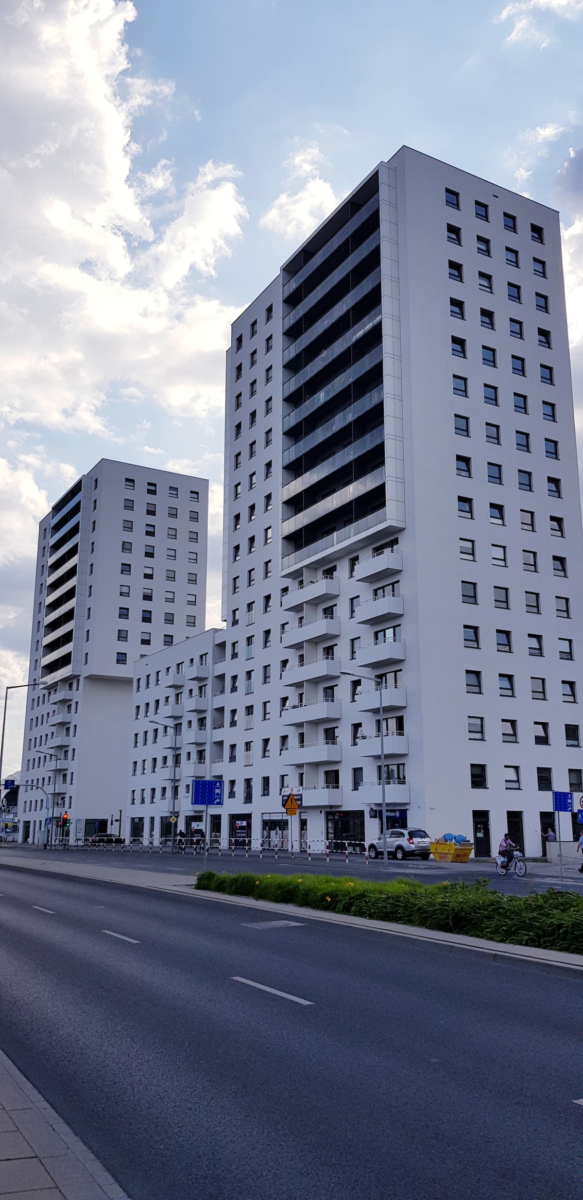 Two modern high-rise buildings are presented, characterized by sleek white facades and numerous windows. Balconies are visible on multiple floors, while the surrounding area includes wide roads and landscaped green spaces, promoting a sense of urban living.