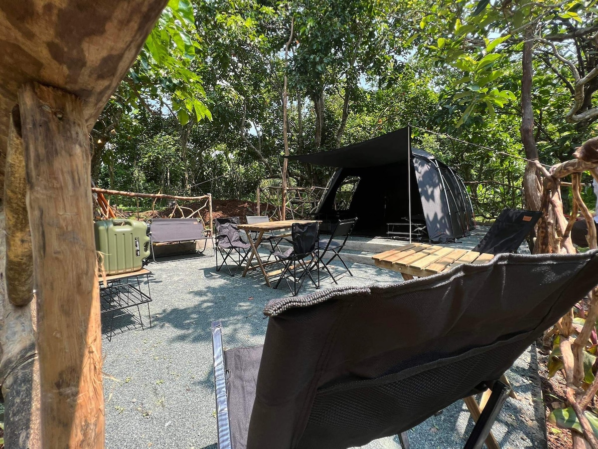 An outdoor camping area is surrounded by lush greenery, featuring a black tent and several camping chairs arranged near a wooden dining table. Various camping equipment is positioned nearby, and a shaded area is provided for relaxation. The ground is covered with gravel for stability.