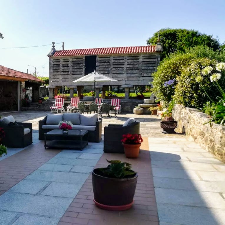 A spacious outdoor area is highlighted by a seating arrangement featuring comfortable sofas and a coffee table. Flower pots with blooming plants add color, while a sun umbrella provides shade. The background includes a traditional hórreo and seating area with striped lounge chairs.