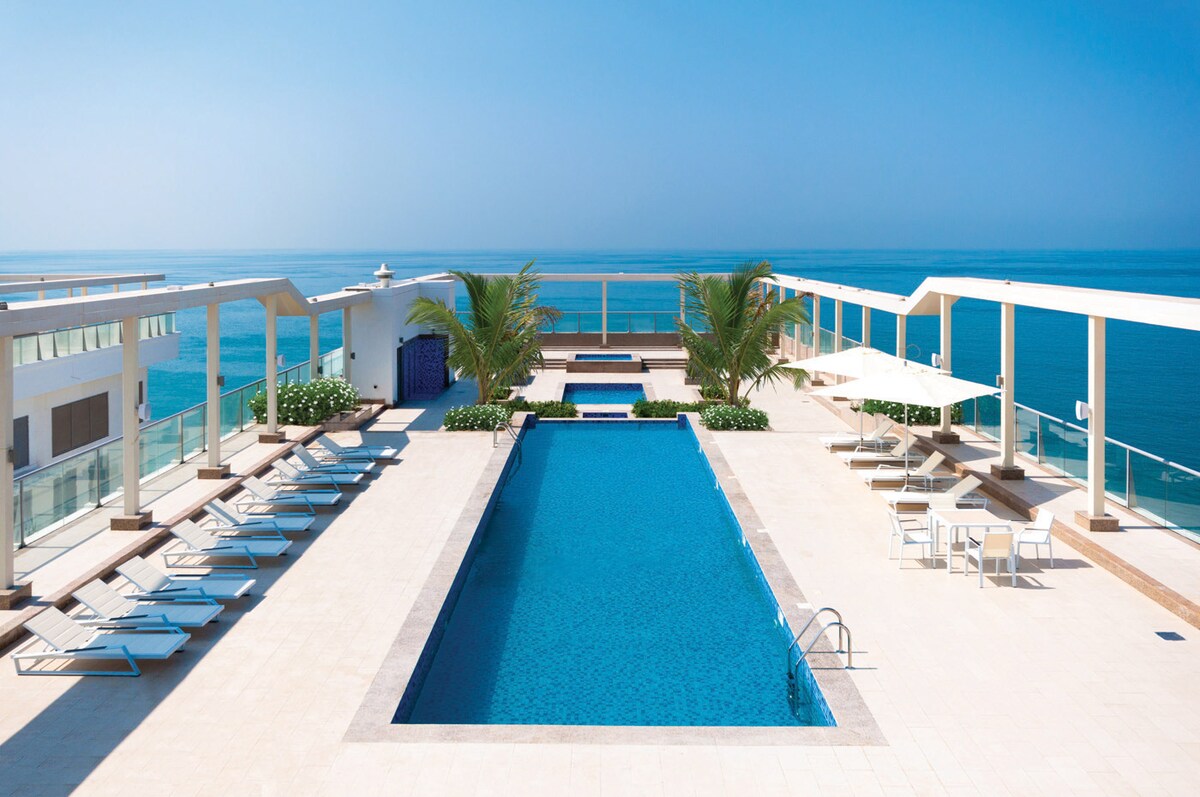 A rooftop area features a large swimming pool surrounded by lounge chairs and tropical plants. Sun loungers are placed along the poolside, and a shaded seating area is visible under an umbrella, with views overlooking the sea and clear blue sky.