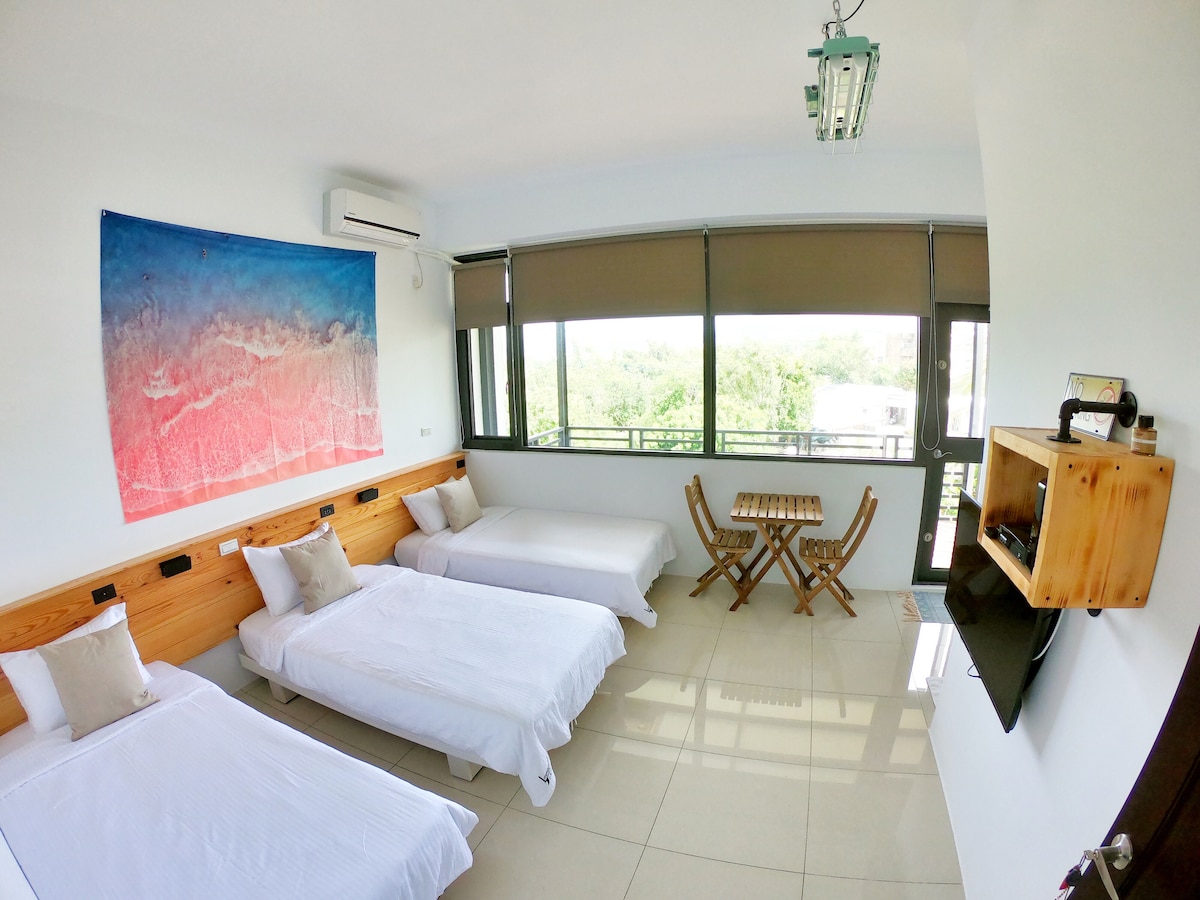 An airy room features three spacious beds arranged with crisp white linens. Natural light floods in through large windows, opening to a balcony view. A wooden table with folding chairs is positioned in the corner, while a wall-mounted TV provides entertainment.