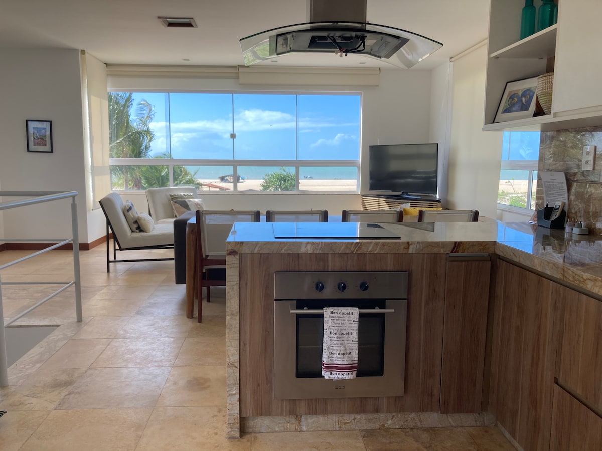 A modern kitchen overlooks a spacious living area, featuring a sleek stove and a large window showcasing ocean views. Soft natural light fills the space, highlighting the minimalist decor and comfortable seating arrangement. A television is positioned for entertainment, with tropical palm trees visible outside.