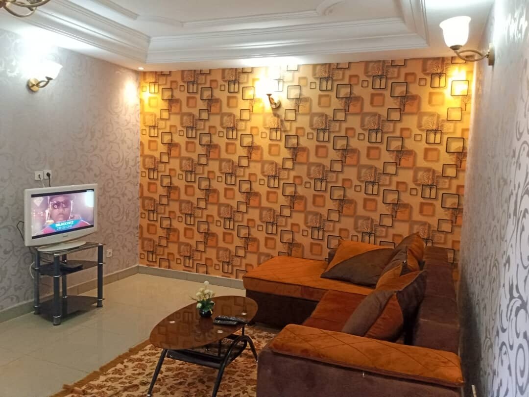 A cozy living area features a sectional sofa adorned with decorative cushions. A coffee table sits in front, and a television is placed on a modern stand. The walls are decorated with a geometric patterned wallpaper, complemented by soft lighting fixtures.