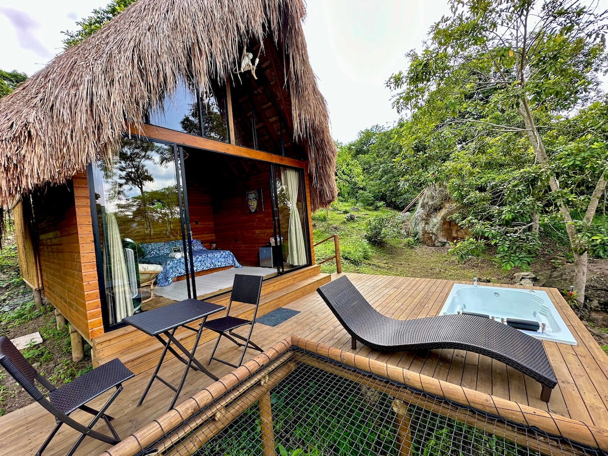A charming wooden cabin features a thatched roof, surrounded by lush greenery. Outdoor seating includes two lounge chairs and a table with four chairs. A private jacuzzi is positioned on the deck, providing a serene space to unwind amid nature.