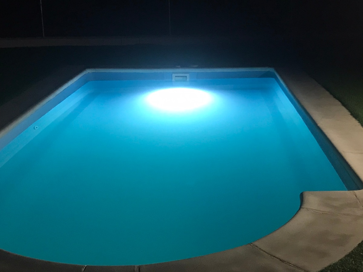 A tranquil swimming pool is illuminated by a soft underwater light, casting a calming glow on the water's surface. The surrounding area is paved, and plush grass borders the pool, creating a serene outdoor environment at night.