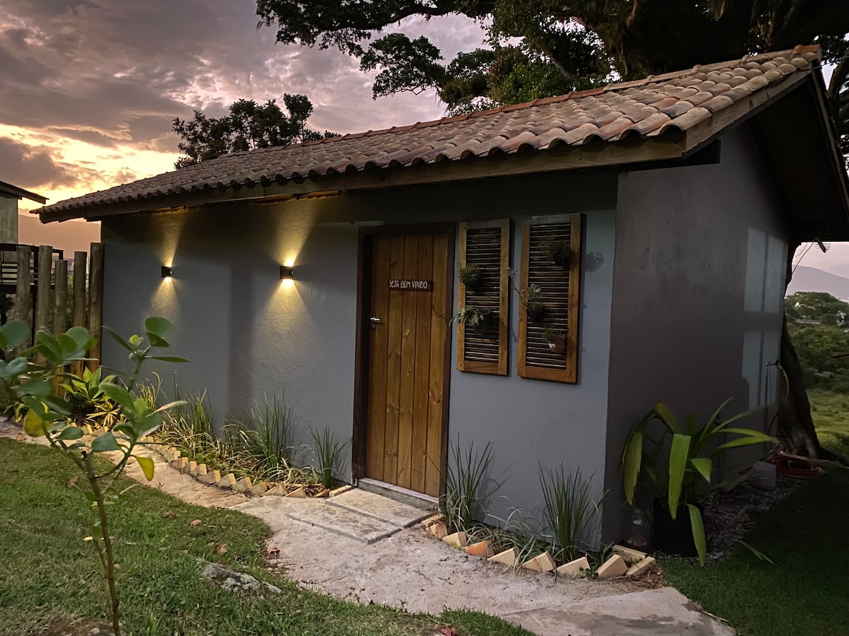 A cozy exterior view of the loft features a wooden door framed by decorative windows. Soft outdoor lighting illuminates the gray walls, complemented by a tidy garden with plants lining the pathway. The scene is set against a backdrop of a dusky sky.