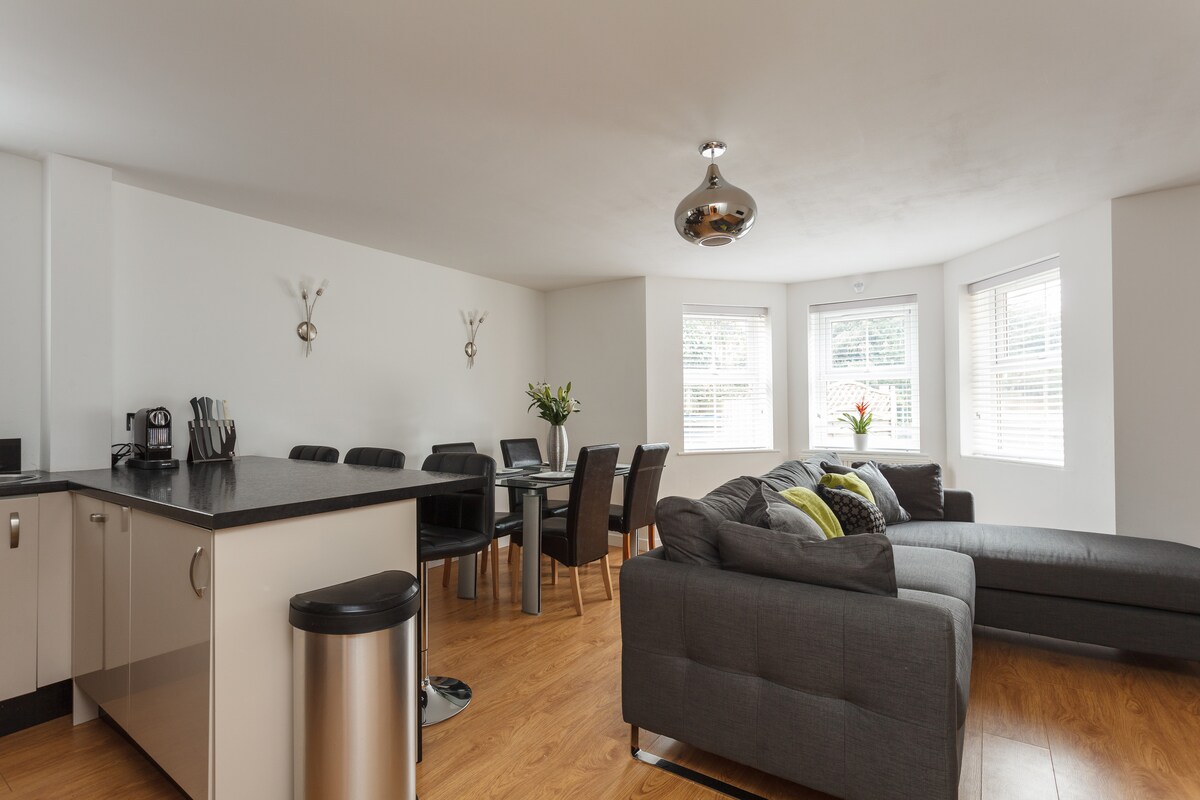 The open-plan living area features a spacious corner sofa in grey, complemented by a coffee table and decorative cushions. A stylish dining table with six black chairs is set near large bay windows, allowing natural light to brighten the space. The modern kitchen is equipped with sleek cabinetry.