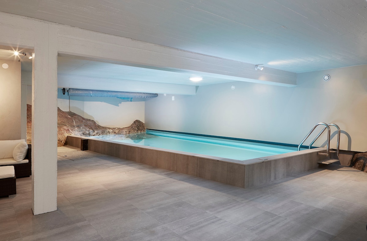 An indoor pool area showcases a spacious, heated swimming pool, framed by smooth stone tiles. Natural light filters through the ceiling, illuminating the environment. A relaxing seating arrangement is positioned to one side, complementing the serene atmosphere of the space.