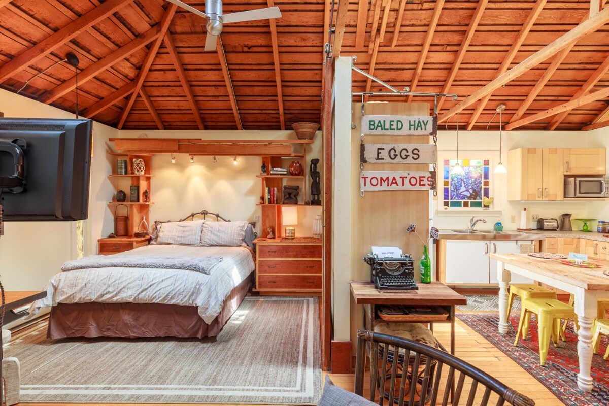 An open living space features a spacious bed with neutral bedding set against wooden cabinetry. A kitchen area with modern appliances is visible, while a dining table with vibrant chairs adds color. A rustic ceiling showcases exposed beams, enhancing the cottage's unique charm.