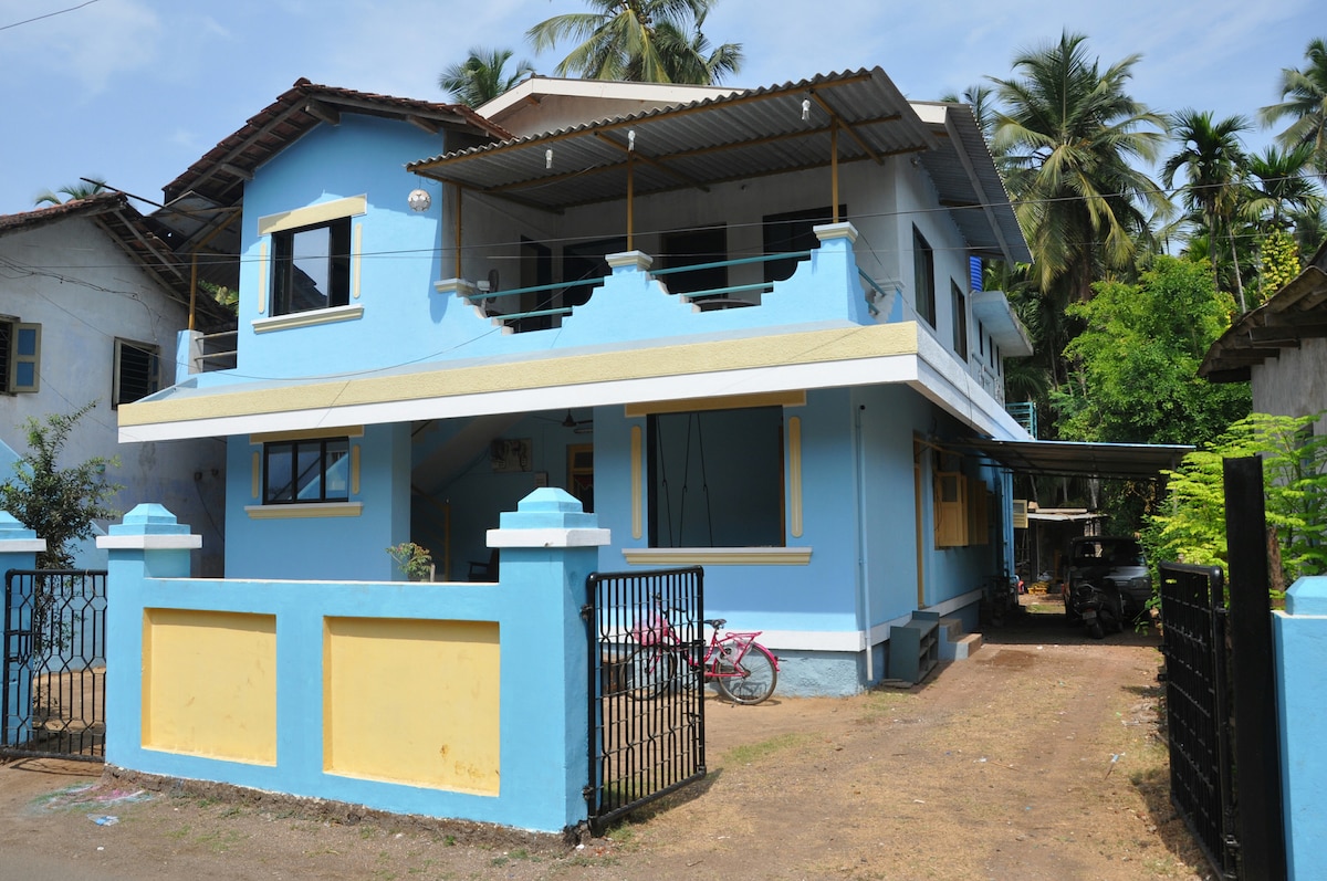 A two-story house with a blue exterior is framed by a low yellow wall. The property features a covered balcony above the entrance and large windows on both floors. Lush greenery surrounds the home, and a pathway leads to the entrance, with a bike visible near the gate.