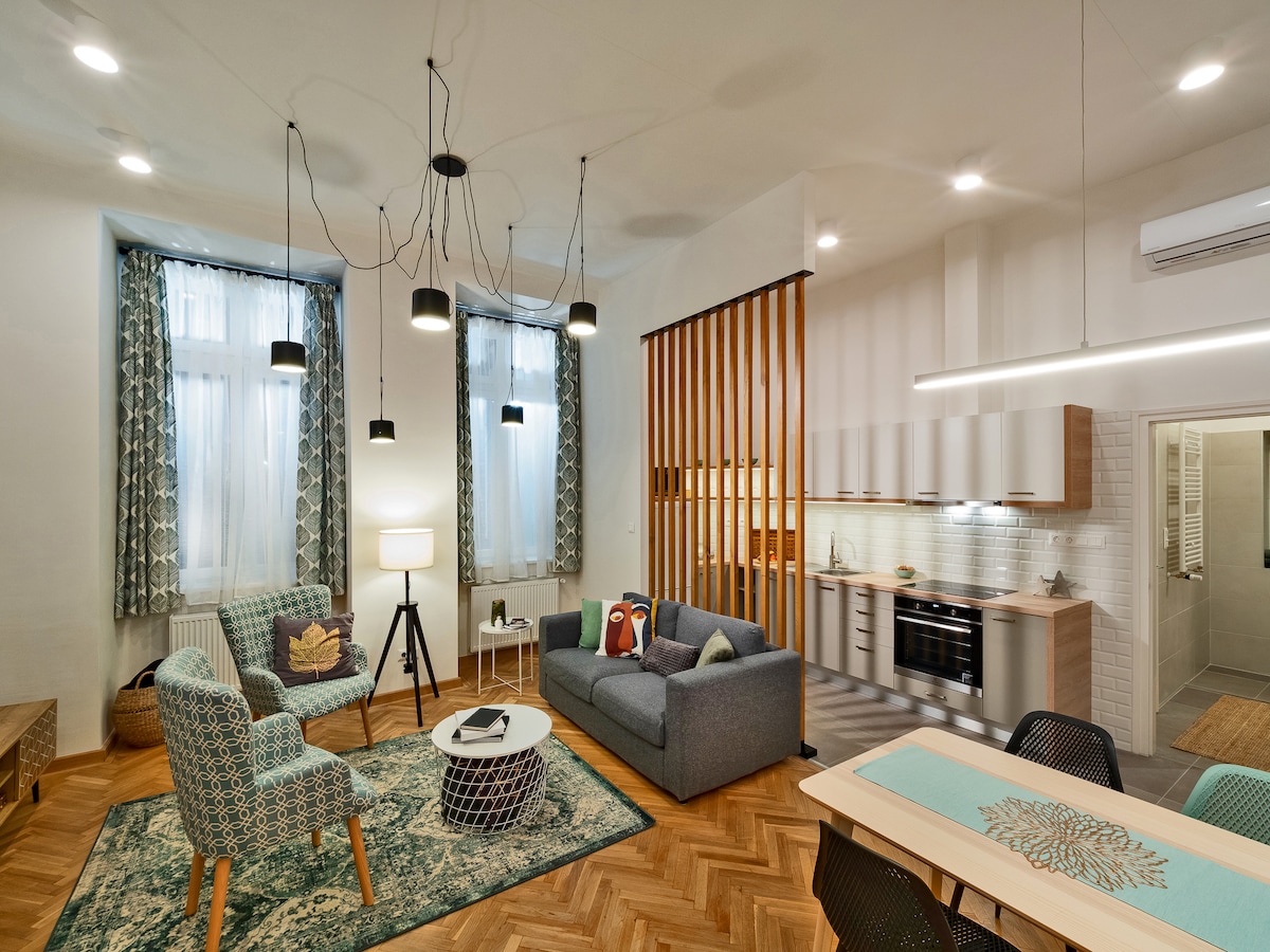 A spacious living area showcases modern furnishings, including a cozy sofa and two patterned chairs. Light streams through tall windows adorned with decorative curtains. The kitchen is visible in the background, featuring sleek cabinetry and modern appliances, while a stylish dining table occupies the area.