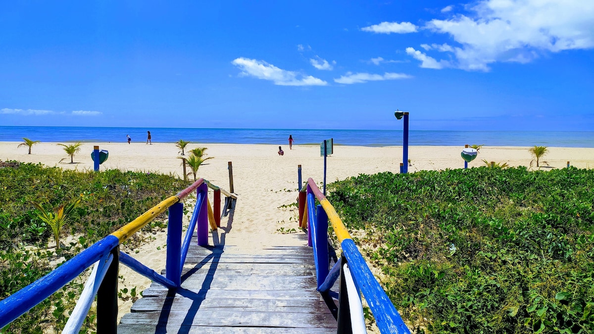 A wooden walkway leads from a lush green area to a sandy beach, framed by low-growing plants. The clear blue sky reflects on the calm ocean, where people can be seen enjoying the water in the distance.