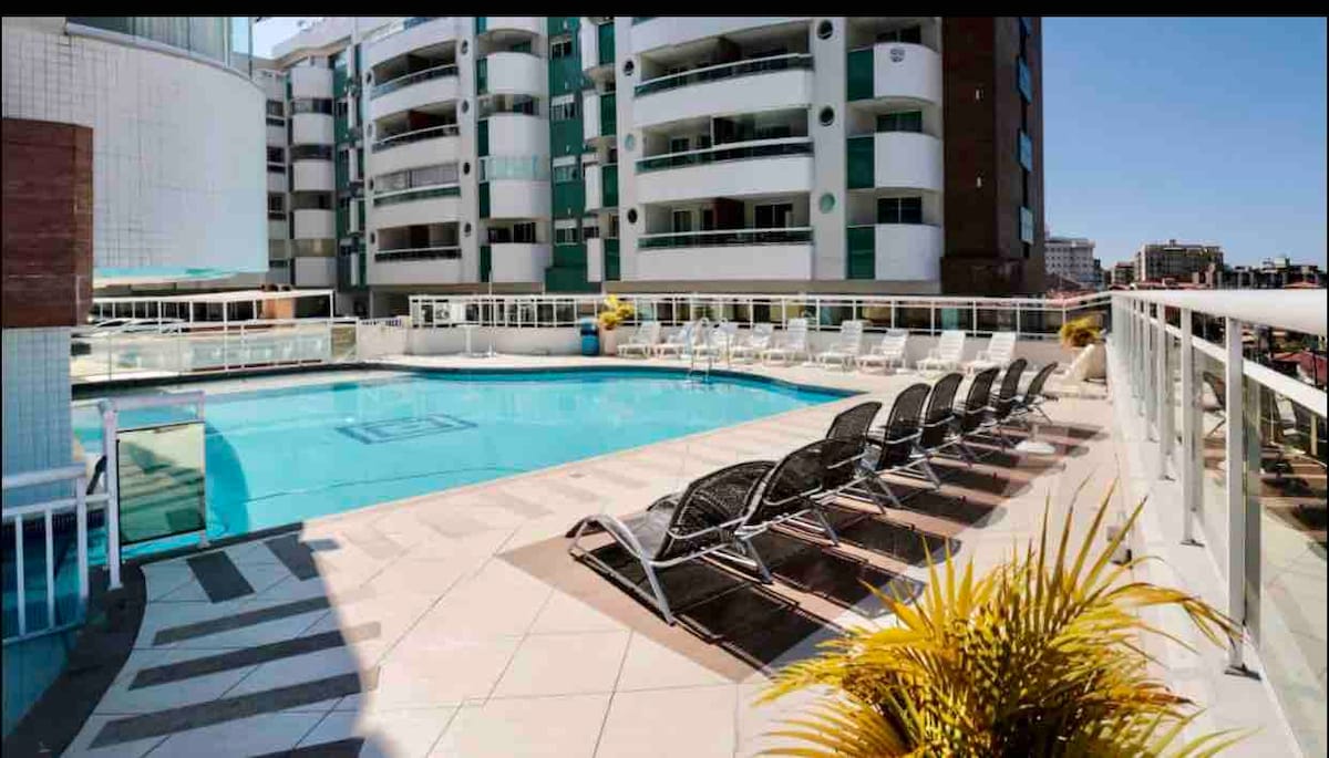 An outdoor pool area is presented with multiple lounge chairs arranged neatly along the sunlit deck. The vibrant blue water of the pool reflects the clear sky, while decorative plants add a touch of greenery to the space.