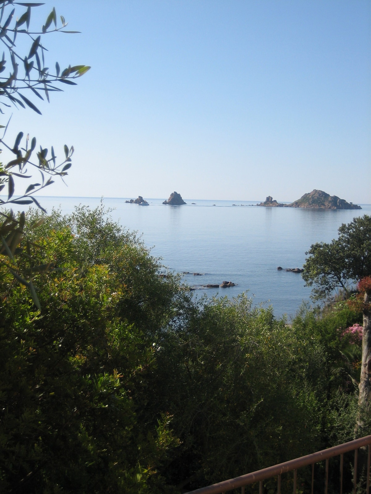 A tranquil view of the sea is framed by lush greenery, showcasing rocky islands rising gently from the calm water. The clear blue sky complements the serene landscape, offering a sense of peace and natural beauty.