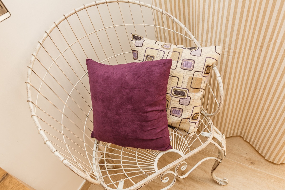 A unique wire chair features two decorative pillows, one in a rich purple hue and another with a geometric pattern, enhancing the inviting seating area. The chair is positioned on a light wooden surface, with soft striped curtains providing a subtle backdrop.