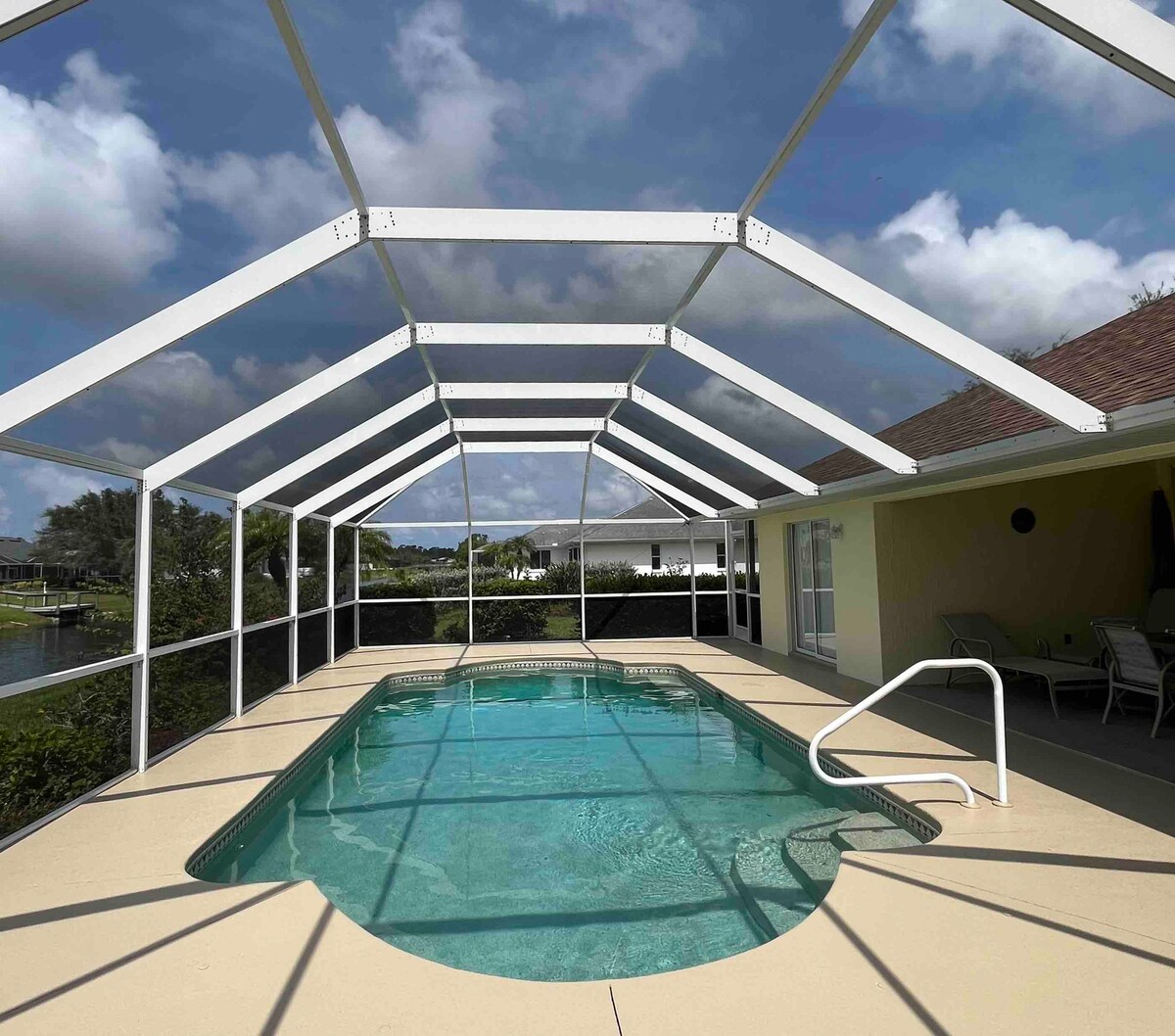 An enclosed pool area is featured, showcasing a private heated pool surrounded by a screened lanai. Visible are pool edges, steps, and a railing. The open sky and clouds create a tranquil ambiance, while lounge chairs are positioned nearby, perfect for relaxation.