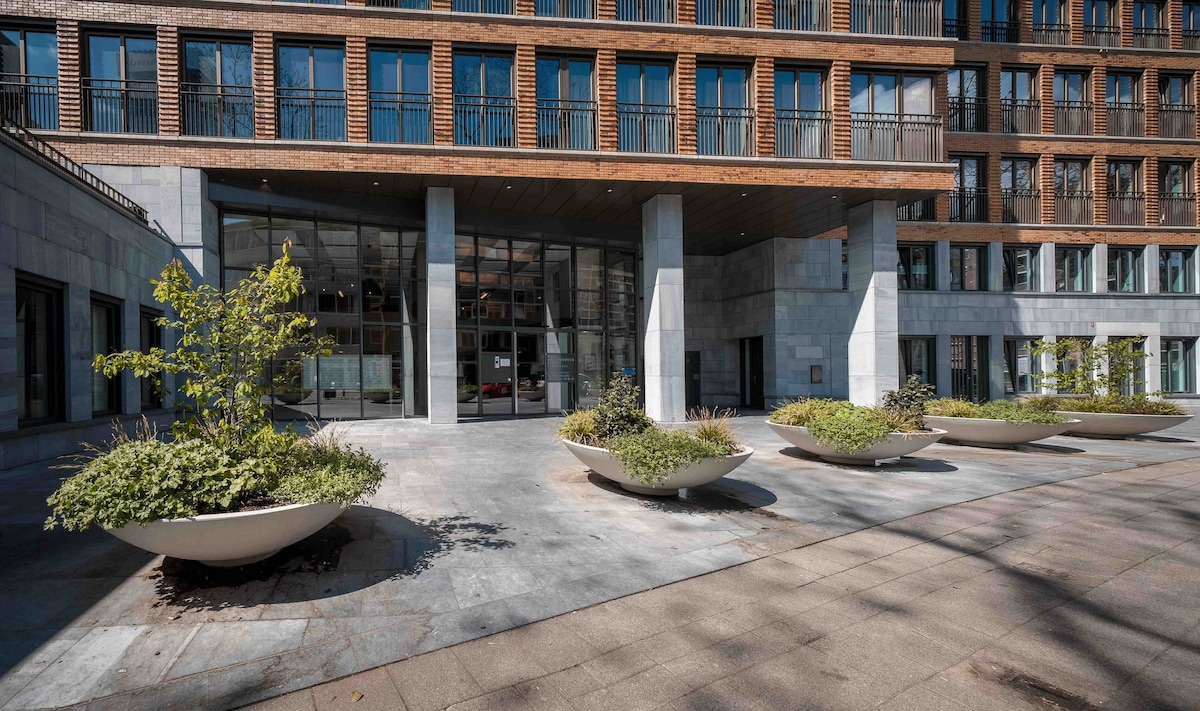 The entrance of a modern building is showcased, featuring large glass doors framed by stone and wooden elements. Planters with greenery are arranged on the smooth surface outside, creating a fresh ambiance. Multiple floor-to-ceiling windows provide a glimpse of the interior.