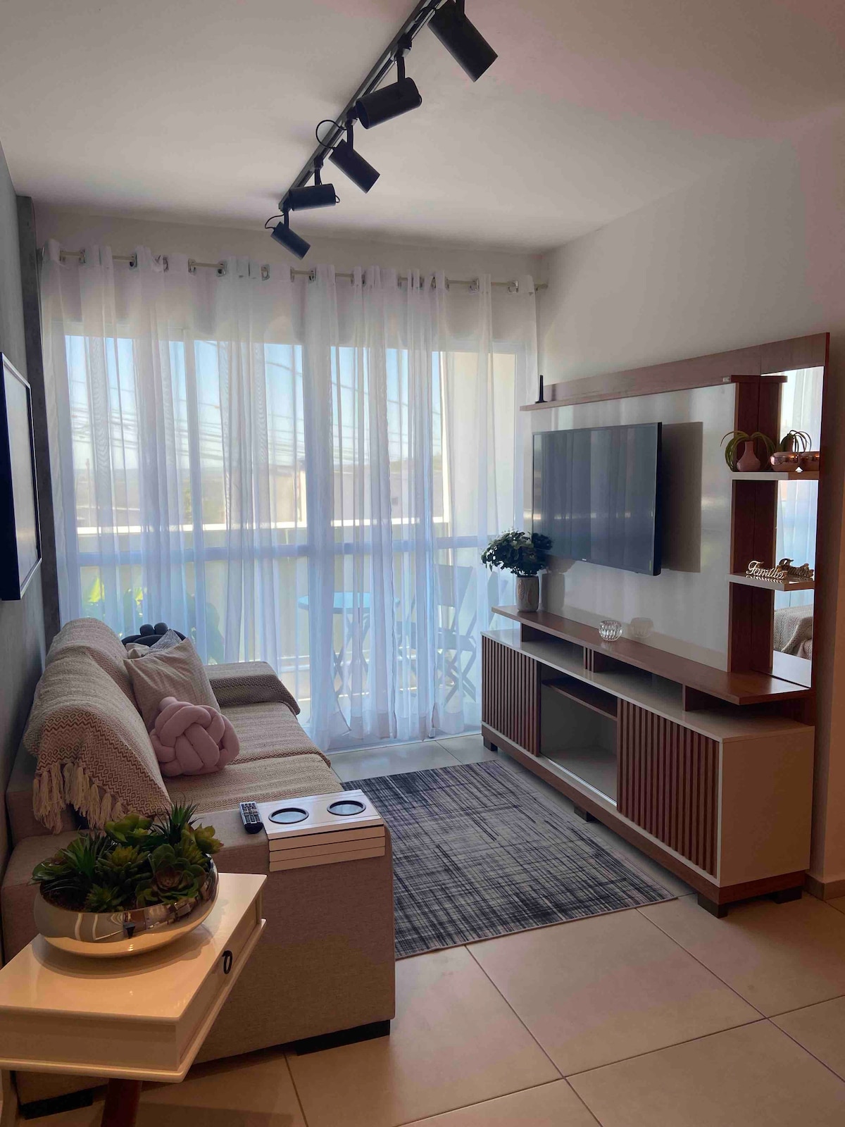 A bright living area is depicted, featuring a light-colored sofa with decorative cushions and a small side table. Natural light filters through sheer curtains, illuminating the space. A wall-mounted television is positioned above a modern console, enhancing the room's functionality.