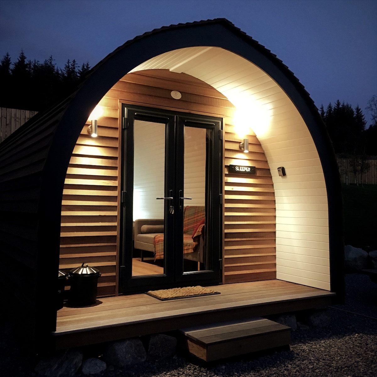An exterior view of a glamping pod is shown at night, featuring an arched design with wooden siding. The entrance is illuminated by wall-mounted lights, highlighting double doors that open to a cozy interior. A welcome mat is placed at the doorway, enhancing the inviting appearance.