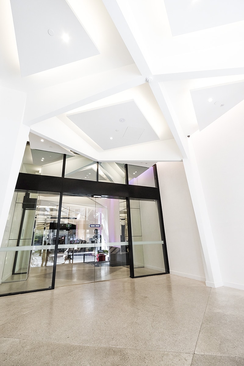 A bright and modern entryway features large glass doors leading into a spacious lobby area. The ceiling showcases a geometric design with recessed lighting, casting a soft glow on the polished concrete floor.