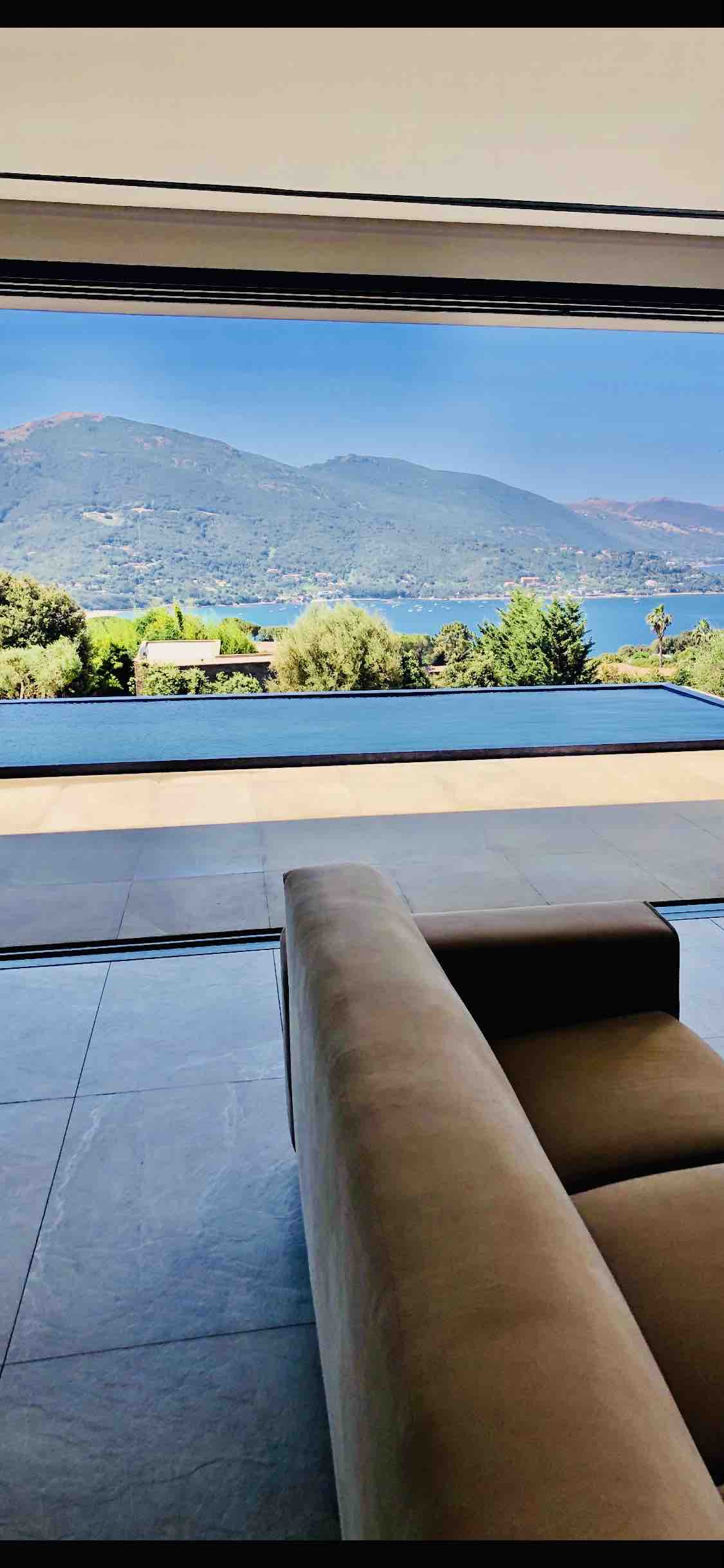 A spacious living area is depicted, featuring a large opening that reveals a view of the mountains and ocean. A modern beige sofa is positioned near the opening, with sleek flooring contributing to the bright and open atmosphere of the space.