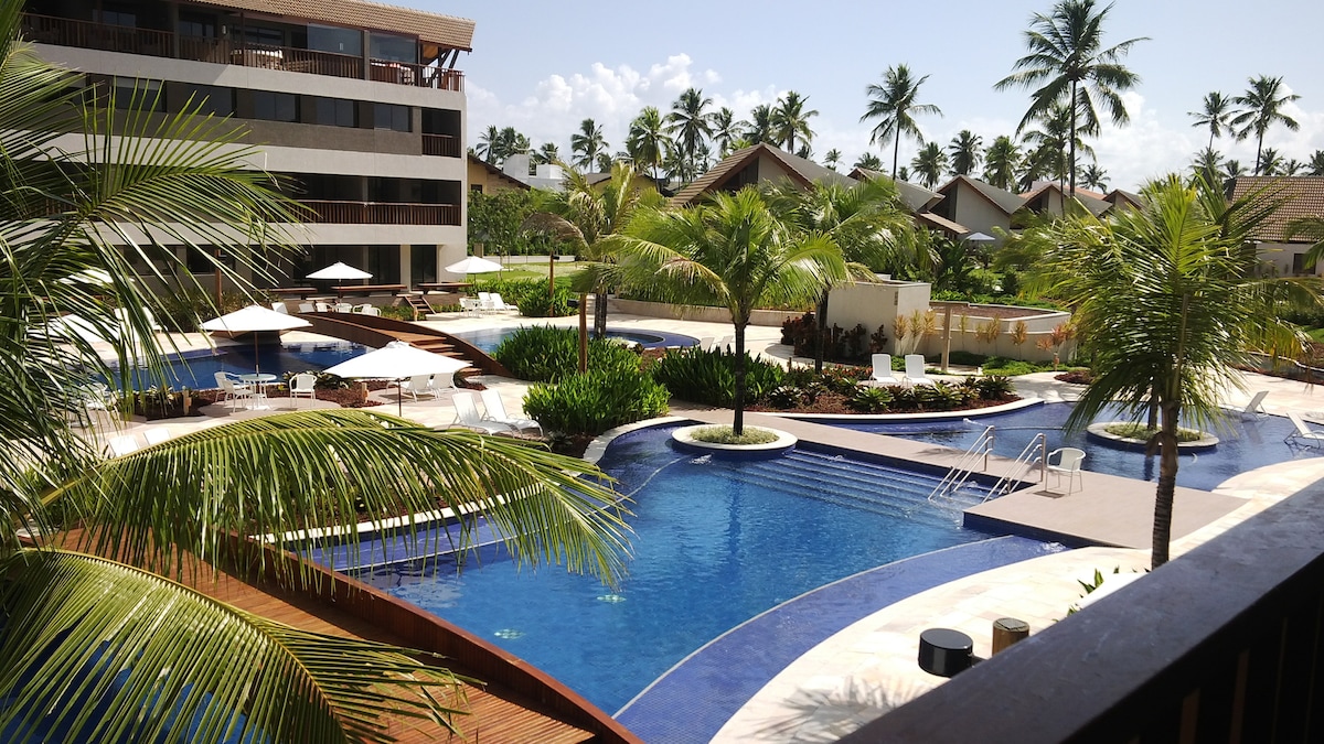 A multi-level pool area is surrounded by palm trees, offering spacious seating areas under umbrellas. Multiple pools are visible, including a larger pool with cascading steps, inviting relaxation in a serene outdoor setting. Lush greenery enhances the tropical ambiance.