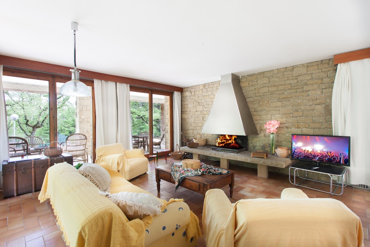The spacious living area features a central fireplace surrounded by comfortable seating with bright yellow throws. Large windows allow natural light to fill the room, and an entertainment unit is visible with a television. Views of the outdoors can be seen through the open windows.