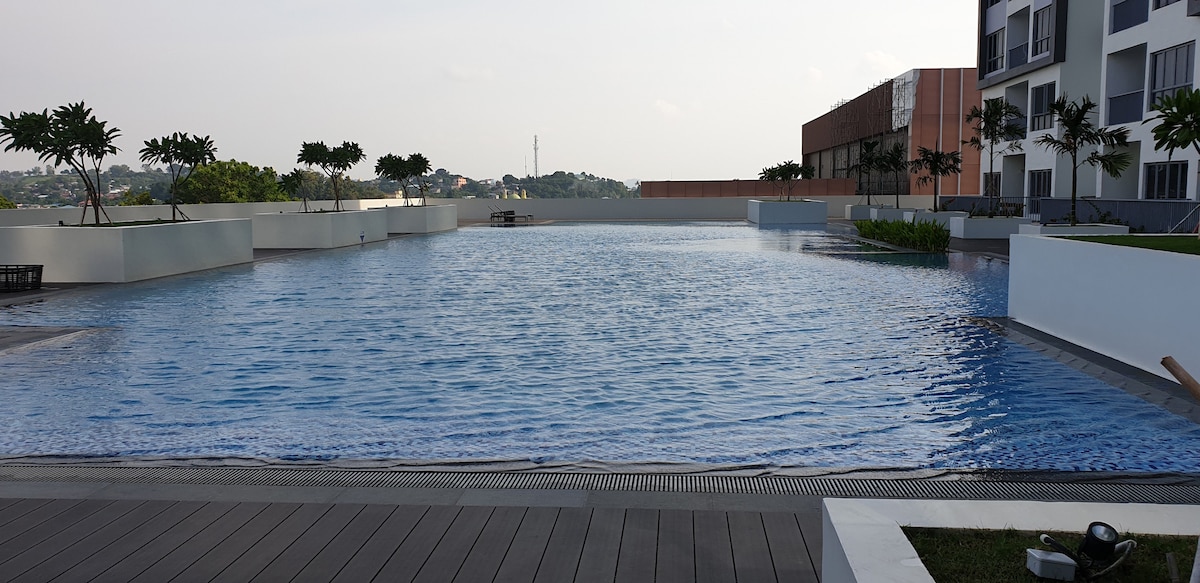 A large infinity pool is visible, reflecting the sky's colors. Surrounding the pool are neatly manicured gardens and trees, providing a serene environment. The modern apartment buildings stand in the background, adding a contemporary touch to the scenery.