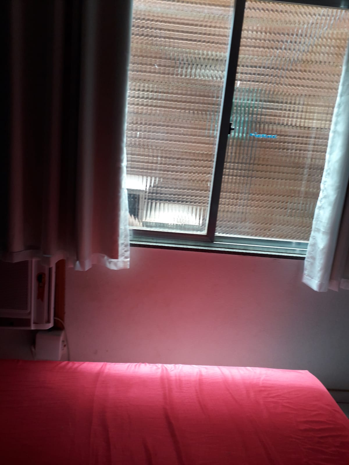 A cozy room is illuminated by soft light filtering through sheer white curtains. A window overlooks a wooden outdoor area, while a spacious bed with a red blanket is positioned below, providing comfort and relaxation.