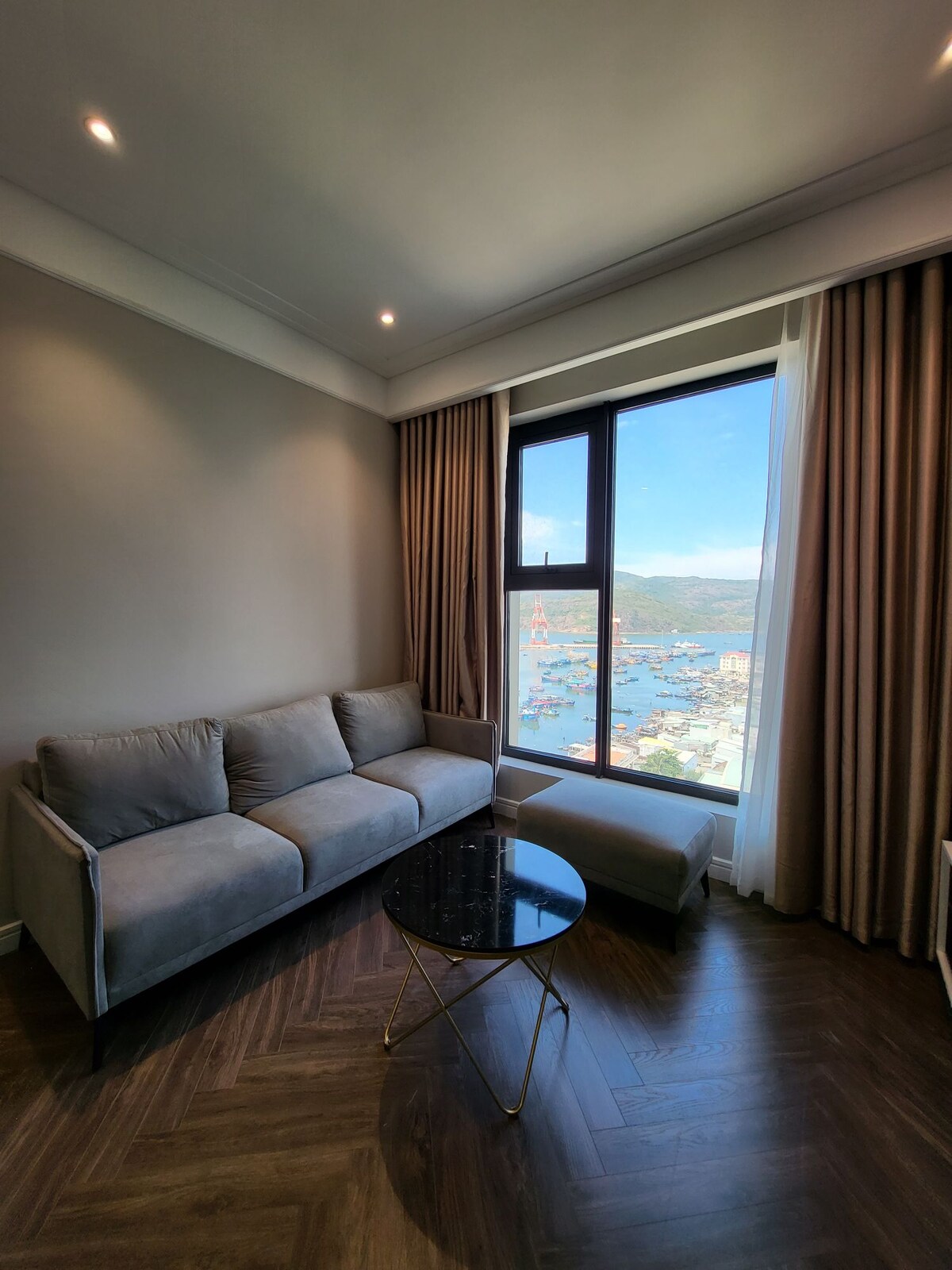 A light-filled corner of the living area features a cozy gray sofa accompanied by an elegant black marble coffee table. Large windows provide stunning views of the harbor, while neutral curtains frame the scenic backdrop, enhancing the overall spacious feel of the room.