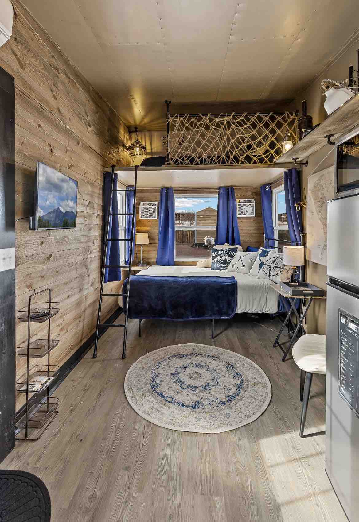 A compact interior features warm wood paneling and a lofted sleeping area. A queen bed is adorned with decorative pillows, while a small kitchenette and refrigerator are visible. Soft blue curtains frame the windows, and a circular rug adds comfort to the wooden floor.