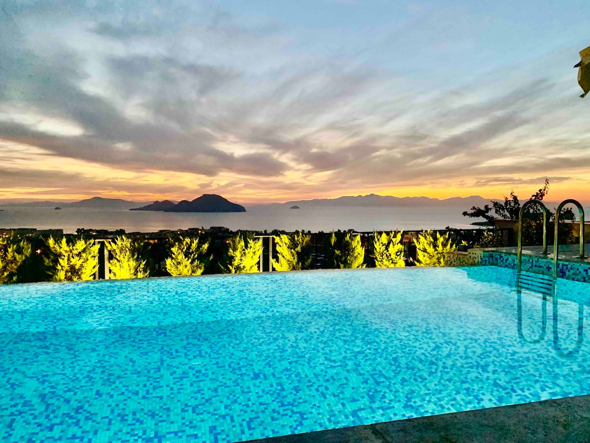 An infinity swimming pool reflects the colors of a sunset, positioned against a backdrop of distant islands and gentle hills. Surrounding greenery is illuminated softly, enhancing the tranquil ambiance of the outdoor space.