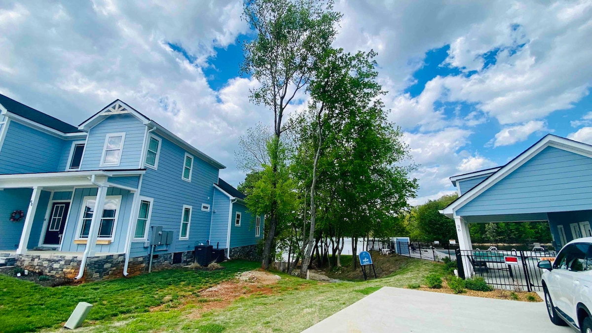 A blue two-story house is depicted alongside a well-kept lawn featuring several trees. The driveway is visible, accommodating up to three vehicles, with the entrance to a gated community on the right. A cloudy sky enhances the serene outdoor environment.