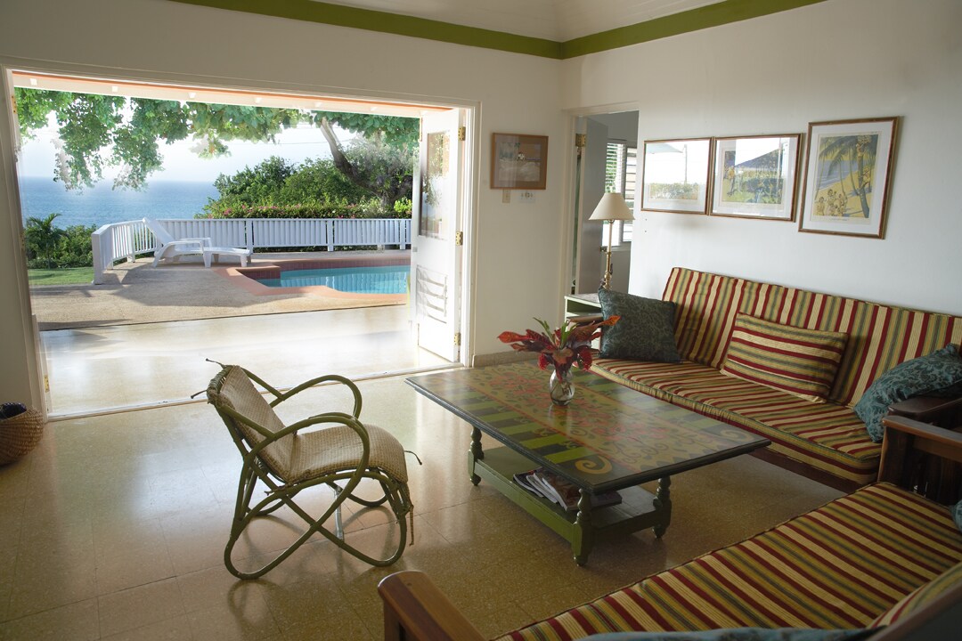 A welcoming living area features a striped sofa set and a glass-topped coffee table adorned with a decorative arrangement. Large sliding doors open to a patio, providing views of the outdoor pool and surrounding greenery.