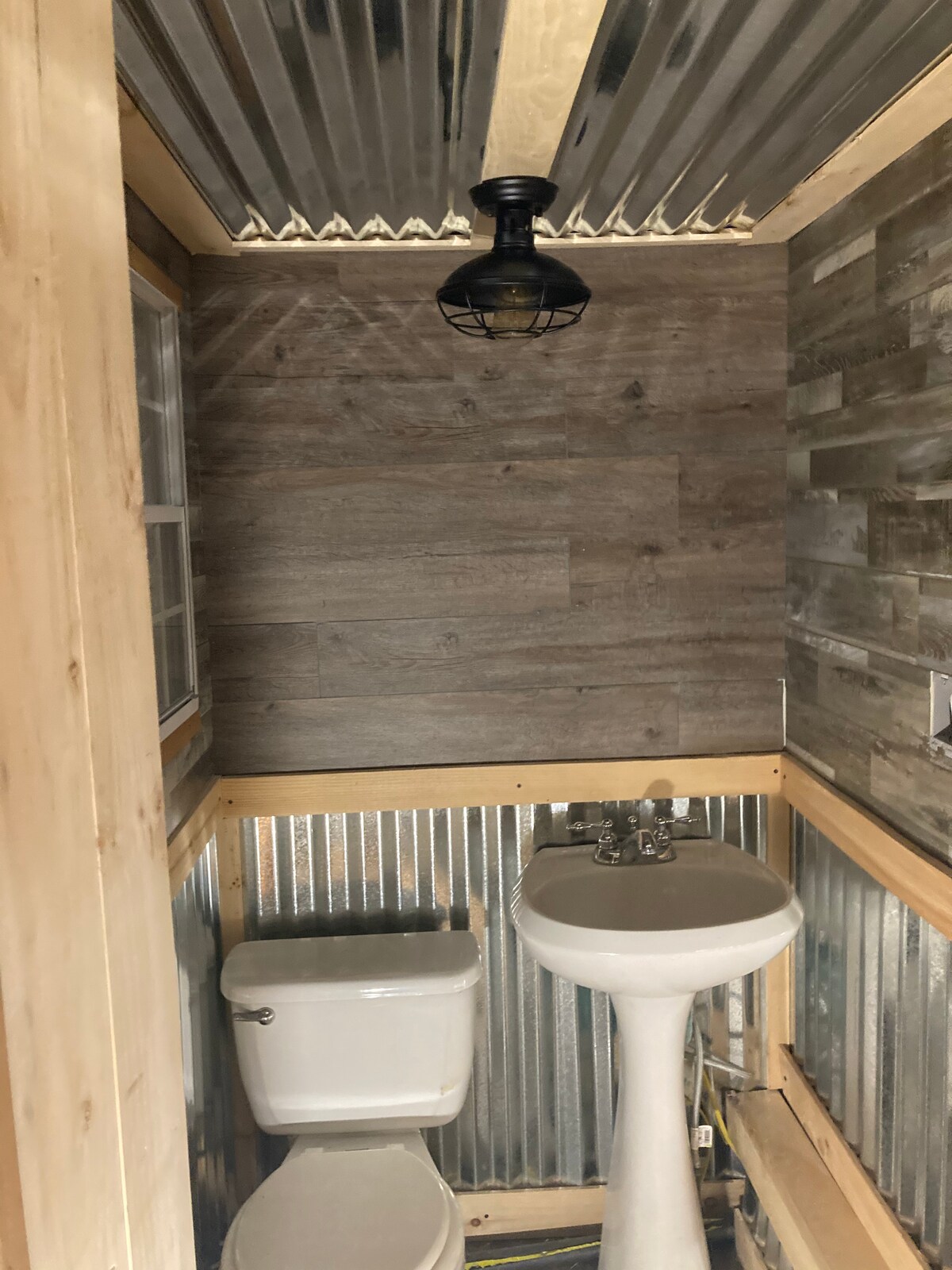A compact bathroom features a combination of rustic elements with corrugated metal and wooden paneling. A pedestal sink and a toilet are situated amidst the warm-toned walls, with a vintage-style light fixture adding character to the space.