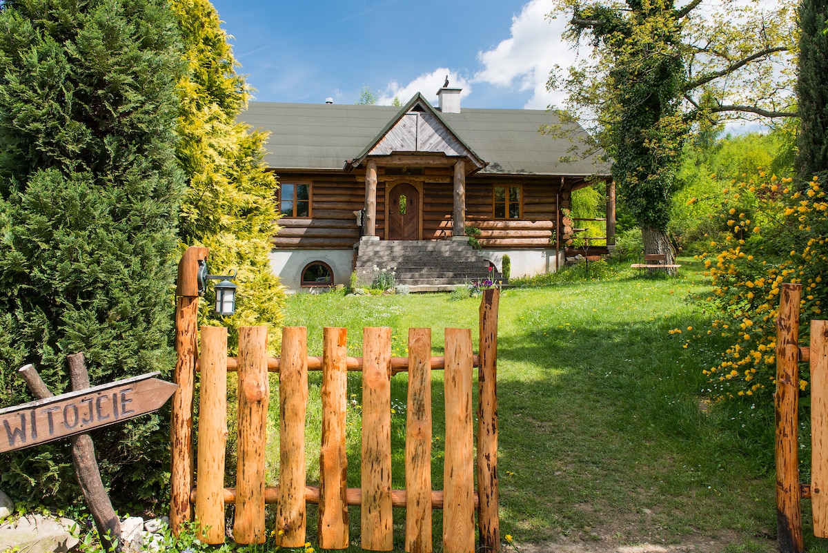 A wooden house with a welcoming front entrance is surrounded by lush greenery. The spacious yard features various trees and blooming bushes, with a wooden fence marking the property. A signpost is visible near the entrance, contributing to the rustic charm of the setting.