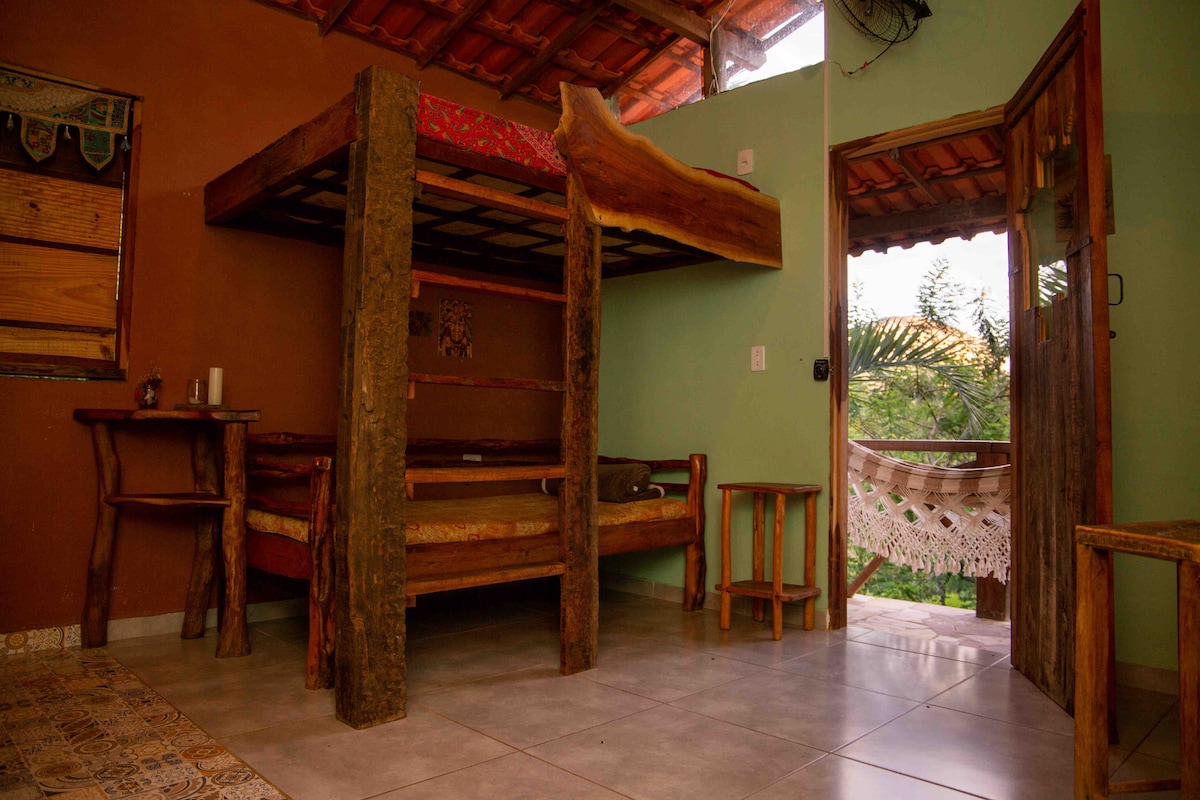 A rustic bedroom features a wooden bunk bed with a bottom sleeping area and a partially visible outdoor balcony. Natural light enters through an open door, illuminating the earthy tones of the walls and the tiled floor. Simple wooden furniture complements the serene setting.