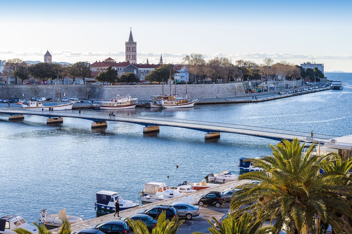 A scenic view of a harbor is captured, featuring boats lined along the water. The shoreline is adorned with palm trees and historic buildings, including a prominent tower. A pedestrian bridge spans the water, connecting both sides, with people leisurely walking across it.