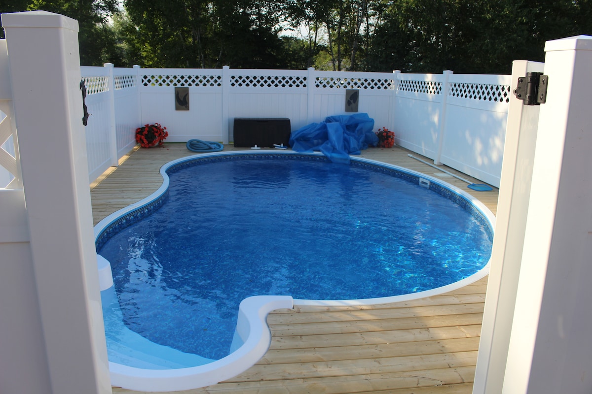 A tranquil pool area is enclosed by a white fenced deck, featuring a clear blue swimming pool with a curved shape. Surrounding plants and seasonal decorations complement the space, while wooden decking adds a warm touch. Pool maintenance equipment is visible nearby.