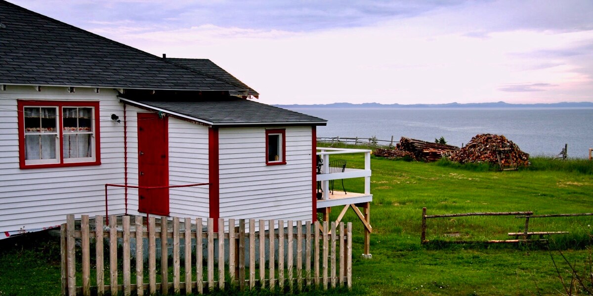 A charming coastal cottage is framed against the backdrop of the ocean. The white exterior features red accents on the door and windows. Grass surrounds the property, and a stacked wood pile is visible in the distance, indicating a serene outdoor setting.