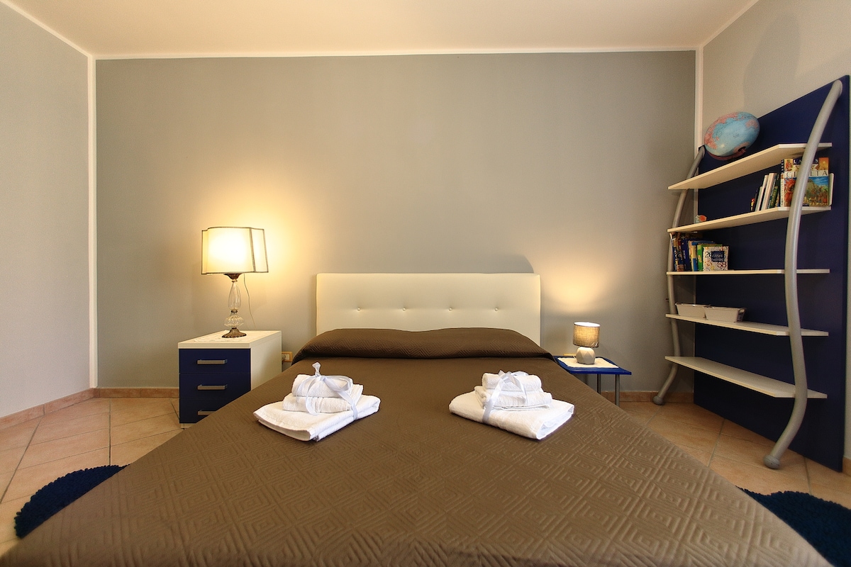 A spacious bedroom features a modern bed with a brown coverlet and neatly arranged towels on top. A bedside table with a lamp is positioned on one side, while a shelf holds books on the opposite wall, illuminated softly by ambient light.