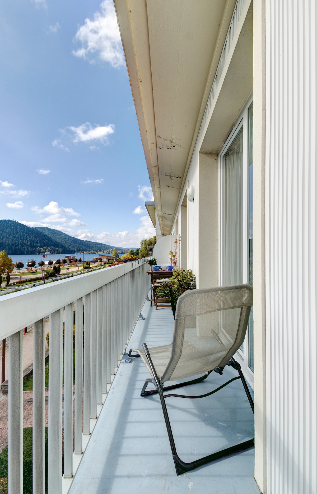 A balcony overlooks a picturesque view of the lake, framed by greenery and distant mountains. A single folding chair provides a comfortable spot to enjoy the outdoors, while the glass railing allows for unobstructed views. The bright sky enhances the serene atmosphere.