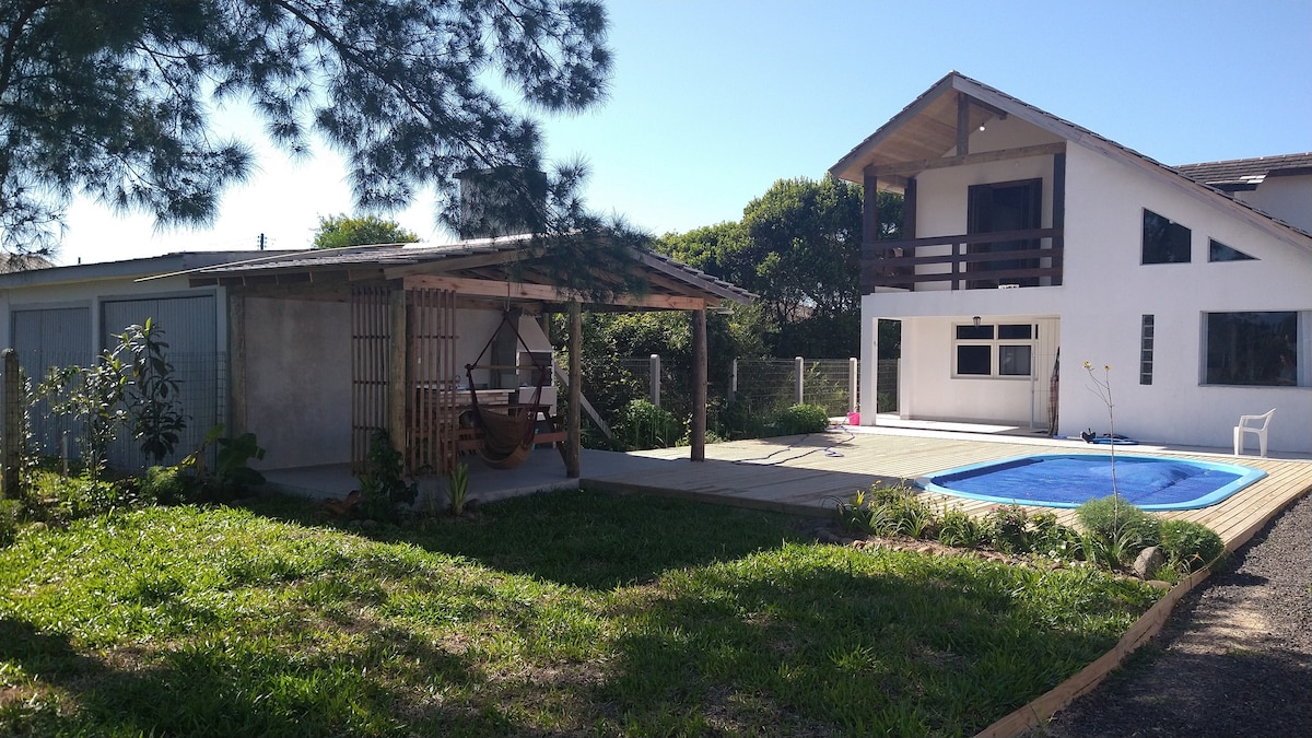 A rustic house with a spacious outdoor area is depicted, featuring a blue swimming pool and a deck. A covered space with a hanging chair provides a relaxing spot. Lush greenery surrounds the property, and the house's large windows allow natural light to fill the interior.