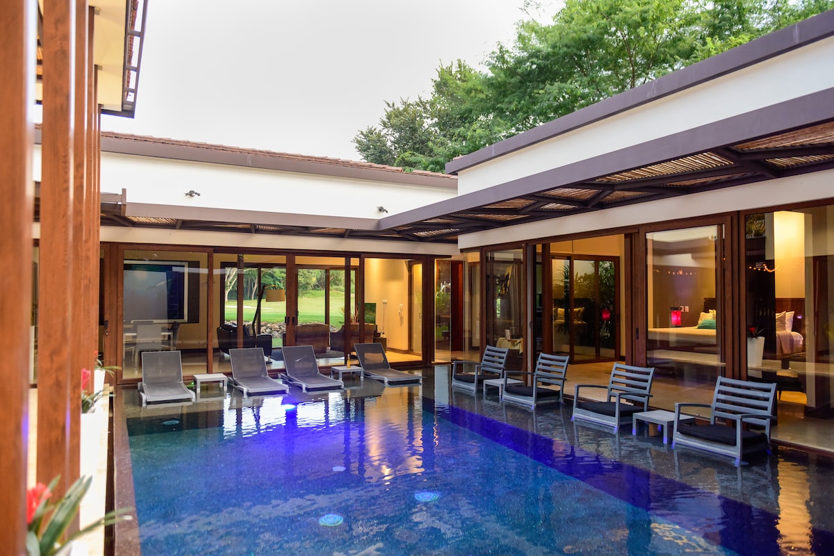 A central swimming pool is surrounded by three separate buildings, with several lounge chairs positioned for relaxation. Floor-to-ceiling windows provide views of the pool and the landscaped grounds. The pool features illuminated water, creating a serene atmosphere in the evening.