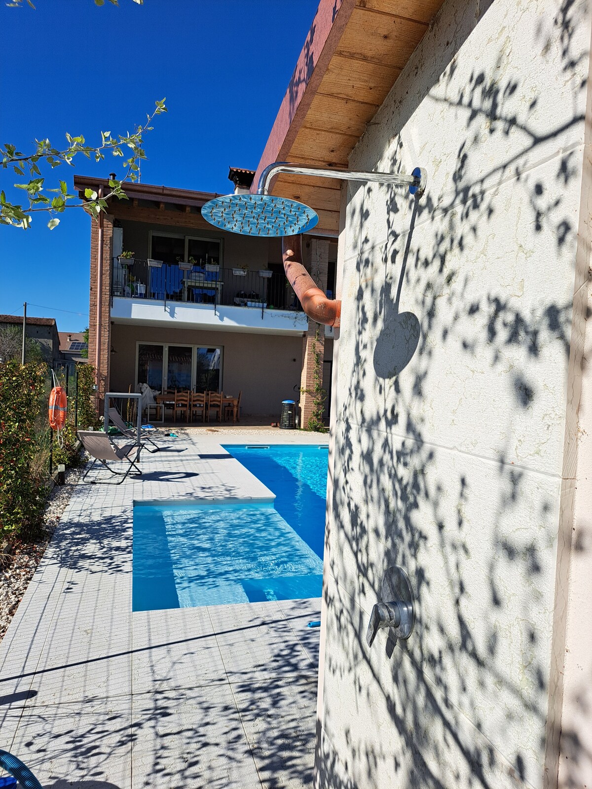An outdoor shower is positioned beside the pool area, featuring a round showerhead and wall-mounted faucet. The pool is visible in the background, surrounded by tiled flooring, with a seating arrangement nearby. Shadows from greenery create a subtle play of light across the scene.