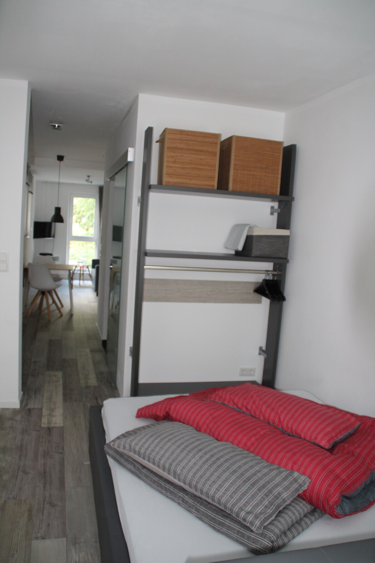 A compact sleeping area features a spacious bed with layered bedding in red and gray stripes. Shelving units are positioned above, holding neatly arranged baskets. The image provides a view into the open corridor leading to the kitchen and living space, offering a glimpse of the apartment's layout.